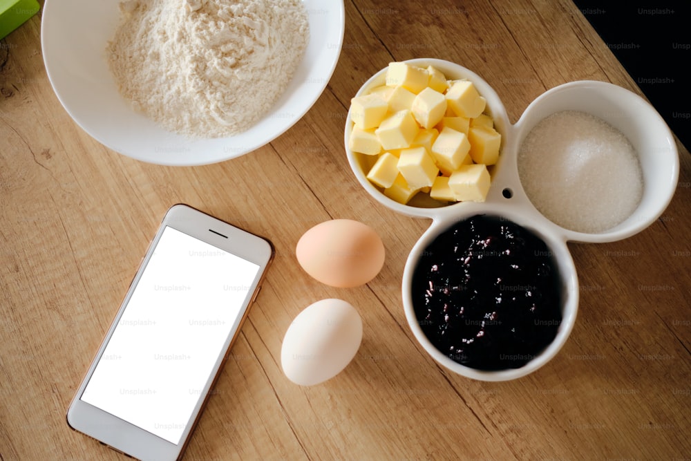 Zutaten für Kuchen auf dem Tisch bereit zum Kochen, während Sie einem Rezept auf dem Smartphone mit weißem Bildschirm folgen. Butter, Edelstein, Eier, Boden