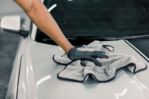 Conceito de detalhamento, lavagem e limpeza do carro. Imagem recortada da mão do trabalhador profissional do sexo masculino em luvas de borracha preta, segurando a microfibra cinza e polindo o capô do carro de carro branco de luxo.