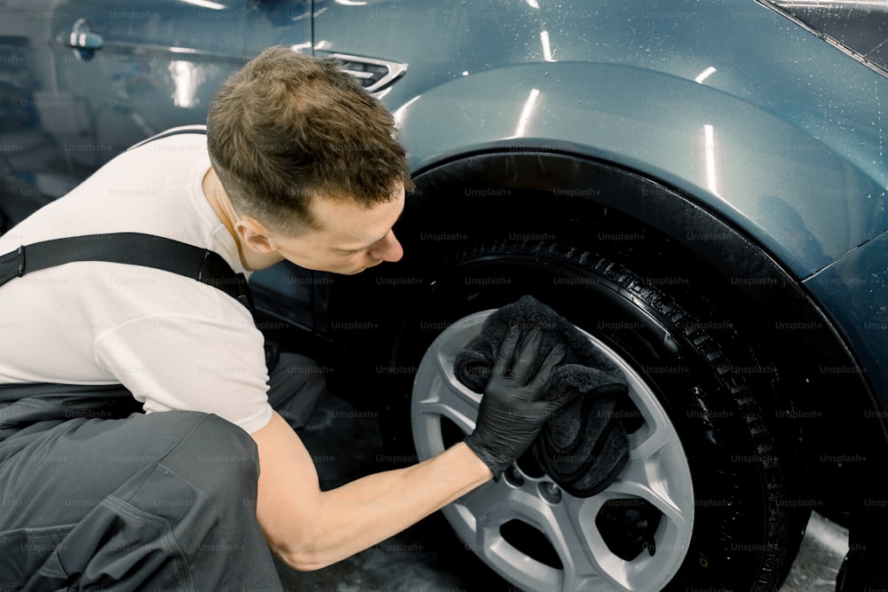 ハンサムな白人男性の洗車作業員が手に黒いマイクロファイバークロスとクリーニングカーホイールをホドリングしている側面図。洗車とディテーリングのコンセプト。