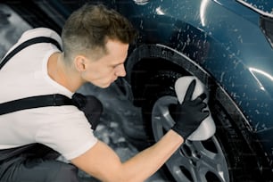 Lavare un'auto a mano. Vista angolare dall'alto del giovane che lava la ruota dell'auto blu moderna con una spugna nel servizio di autolavaggio.