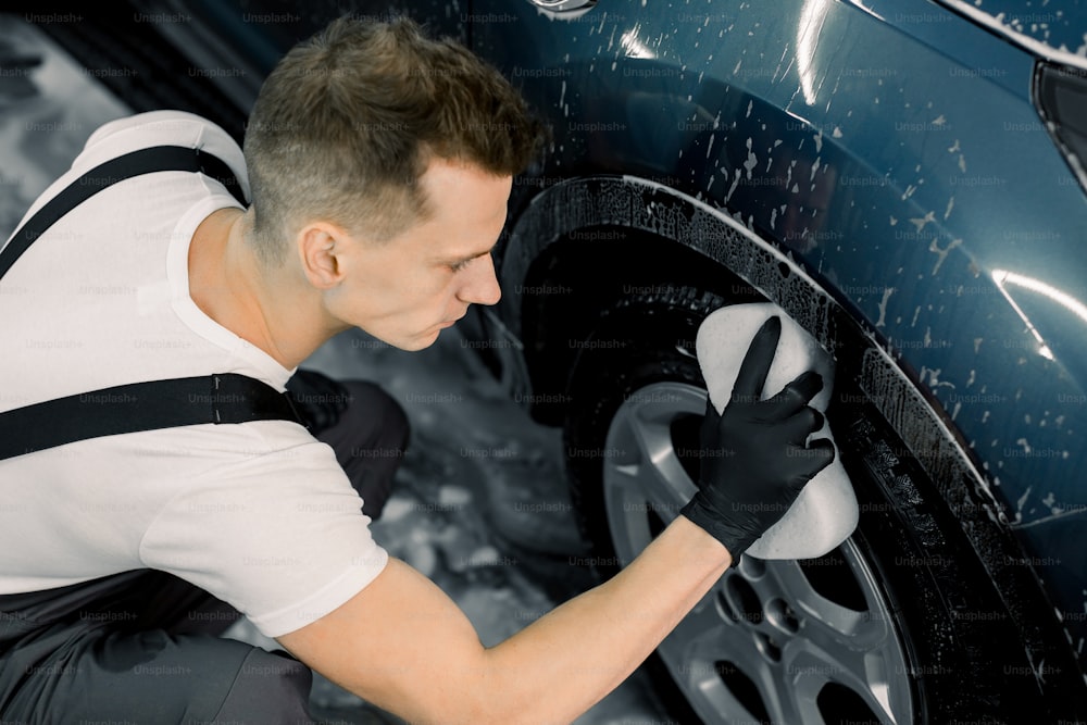 Washing a car by hand. Top angle view of young man washing the wheel of modern blue car with a sponge in carwash service.