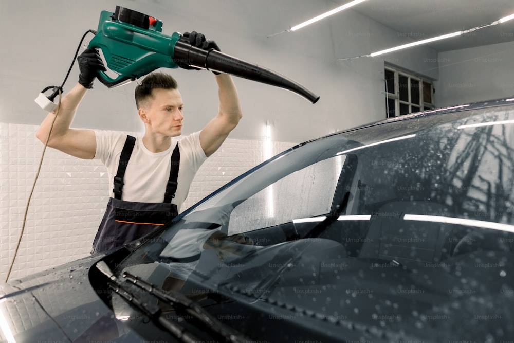 Car washing and detailing concept. Professional young male car service worker holding special machine and drying wet car after washing with water