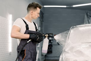 Car wash with flowing water and foam. Side view of young car wash service male employee holding high pressure spray with cleaning foam. Carwash. Washing machine at the station.