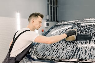 Auto waschen und mit Shampoo und Schwamm reinigen. Junger gutaussehender Arbeiter, der seifige Autowindschutzscheibe mit Schwamm auf einer Autowaschanlage wäscht