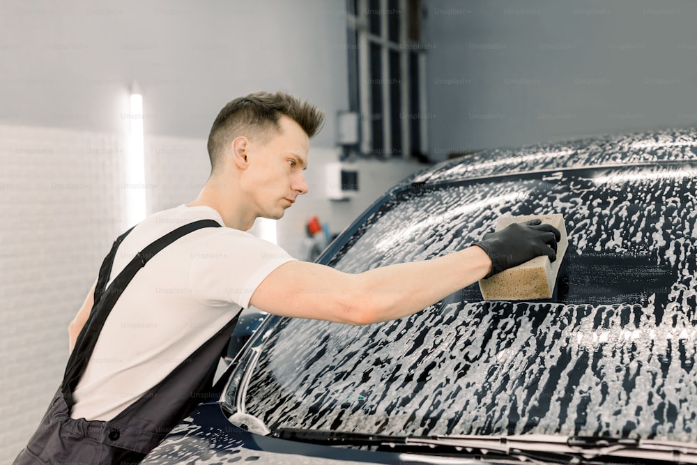 Car wash and clean with shampoo and sponge. Young handsome man worker washing soapy car windshield with sponge on a car wash