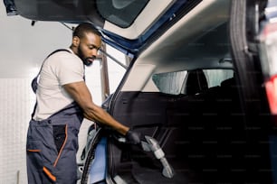 Trabajador masculino africano con camiseta blanca y mono gris, aspirando el interior del automóvil, maletero con aspiradora húmeda, método de extracción profesional. Aspiradora de coche mojado.