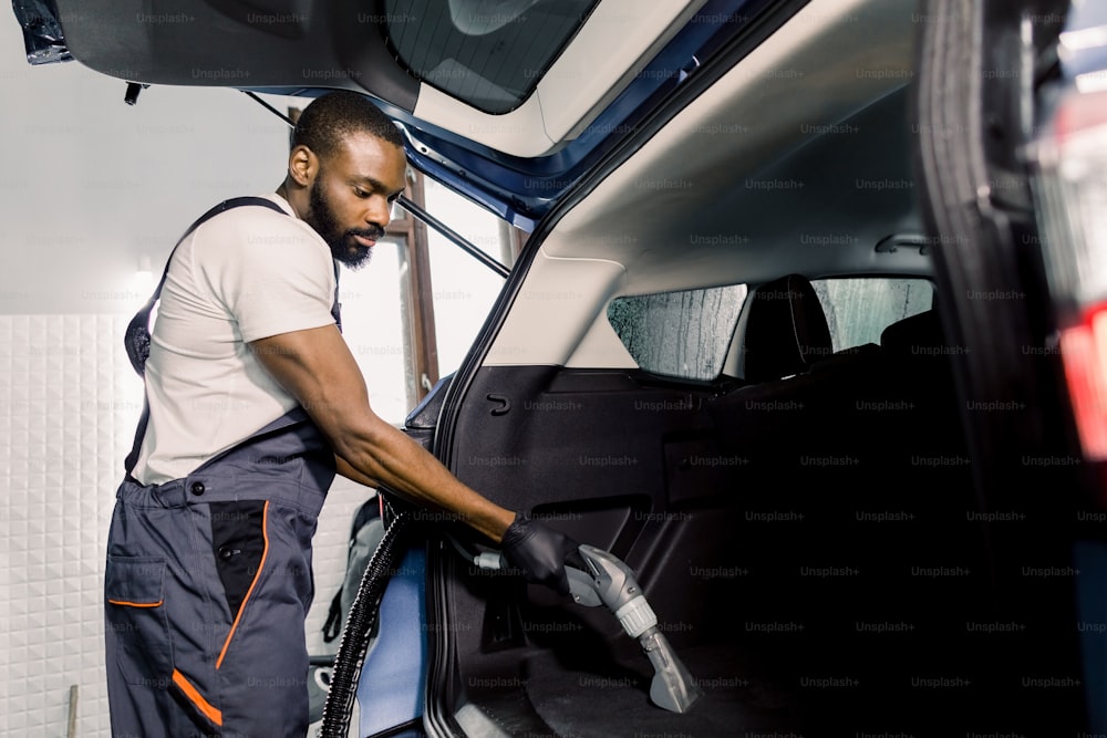 Trabalhador africano vestindo camiseta branca e macacão cinza, aspirando o interior do carro, porta-malas com aspirador de pó molhado, método de extração profissional. Limpeza a vácuo de carro molhado.