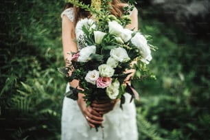 stylish boho bride holding  rustic bouquet of amazing flowers and herbs on background of rocks in mountains