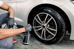 Lavaggio di un'auto a mano, car detailing. Immagine ritagliata di mani di giovane uomo in guanti protettivi che spruzzano una soluzione detergente speciale sul bordo di una moderna auto sportiva bianca con servizio di autolavaggio.