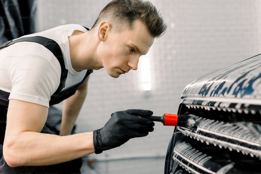 Close up photo of young handsome man in black protective gloves cleaning radiator grille in foam with special brush. Carwash and detailing. Washing machine at the station. Car washing concept
