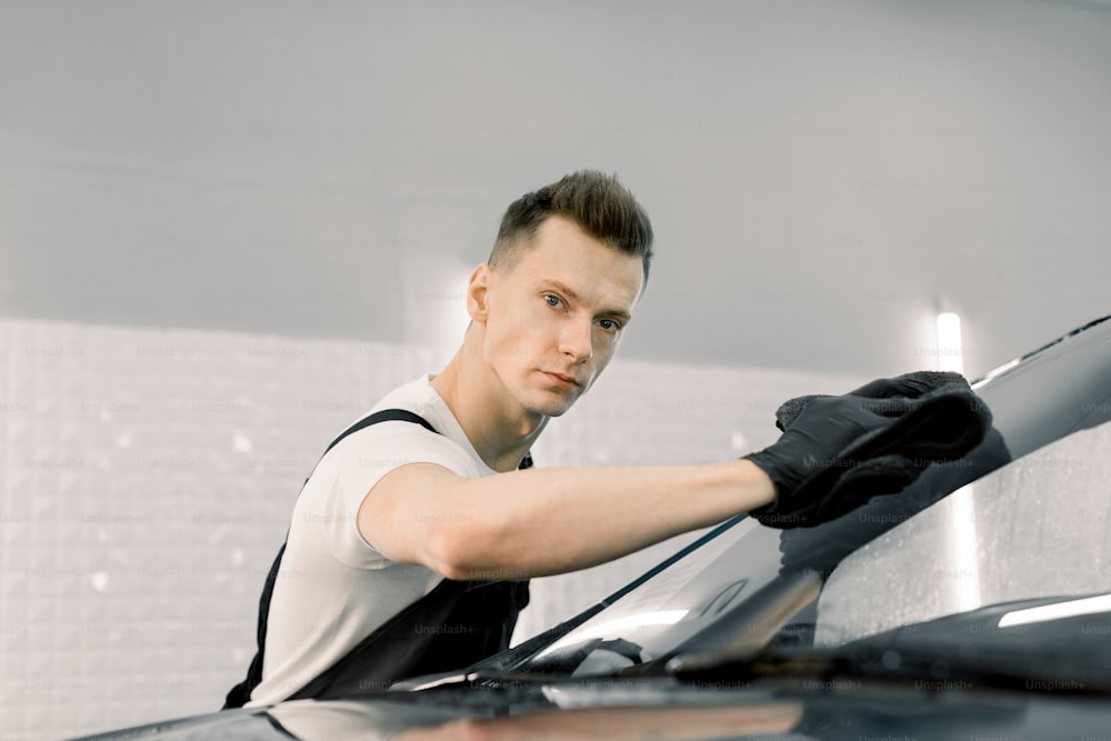 Autoservice männlicher Arbeiter in Schutzhandschuhen und einheitlicher Reinigung Autowindschutzscheibe mit Mikrofasertuch und Blick in die Kamera. Fahrzeugdetaillierung und Valeting-Konzept.