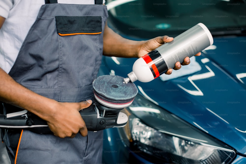 Cropped image of African man, auto service worker, wearing white t-shirt and gray overalls, putting special polish wax or cream on the orbital polisher to polish the blue car behind.