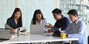 モダンな会議室を背景に、白いミーティングデスクに一緒に座りながら、コンピューター、ラップトップ、タブレットで会議をしているビジネスマンの写真。