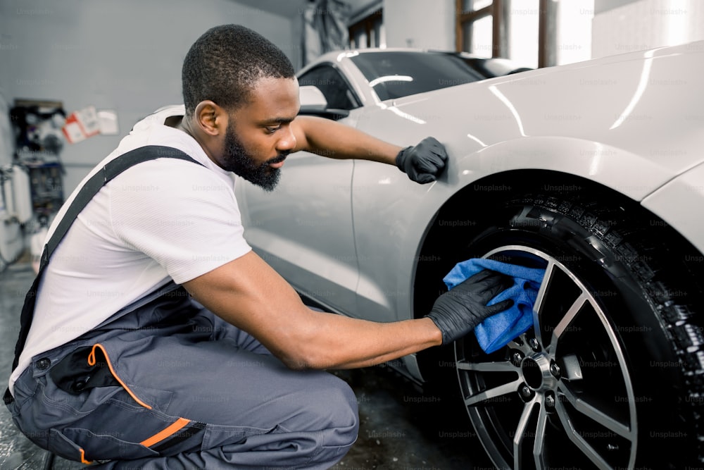 洗車と詳細写真。保護オーバーオールとゴム手袋を着用し、マイクロファイバークロスと特別なクリーナーを使用して、洗車で車の合金ホイールを洗うアフリカの男性労働者。