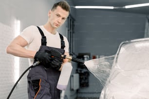 Retrato de perto do trabalhador da lavagem de carros, jovem hansome em macacão de proteção, limpando o automóvel com jato de água de alta pressão na lavagem do carro, pulverizando a espuma de limpeza.