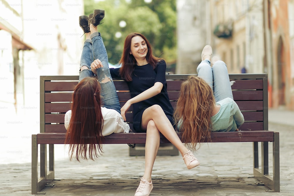 Mujeres felices con estilo hipsters sonriendo y sentadas en el banco en la calle de la ciudad de Europa, divirtiéndose al revés, concepto de amistad