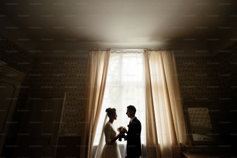 mariée et le marié de luxe heureux debout à la lumière de la fenêtre dans la chambre riche, moment tendre