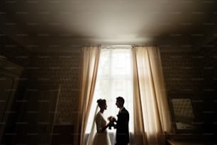 Novia y novio de lujo felices de pie en la luz de la ventana en la habitación rica, momento tierno