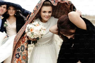 花婿付添人と花嫁介添人とスカーフの下に花束を持つ幸せな花嫁が楽しんでいる、屋外で雨が降る、陽気な瞬間