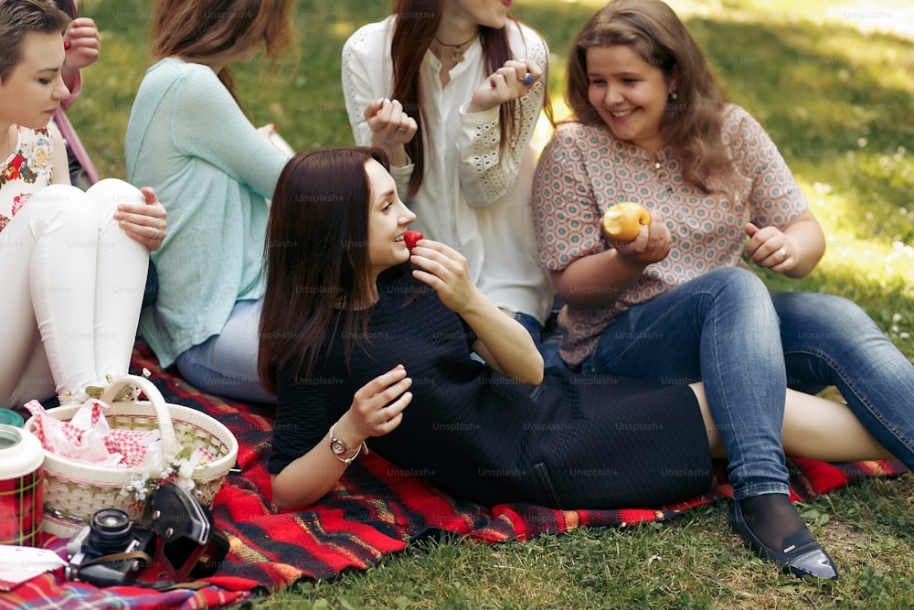 Gruppo felice alla moda di donne che mangiano frutta e si divertono sorridendo durante il picnic, momenti gioiosi nel parco estivo