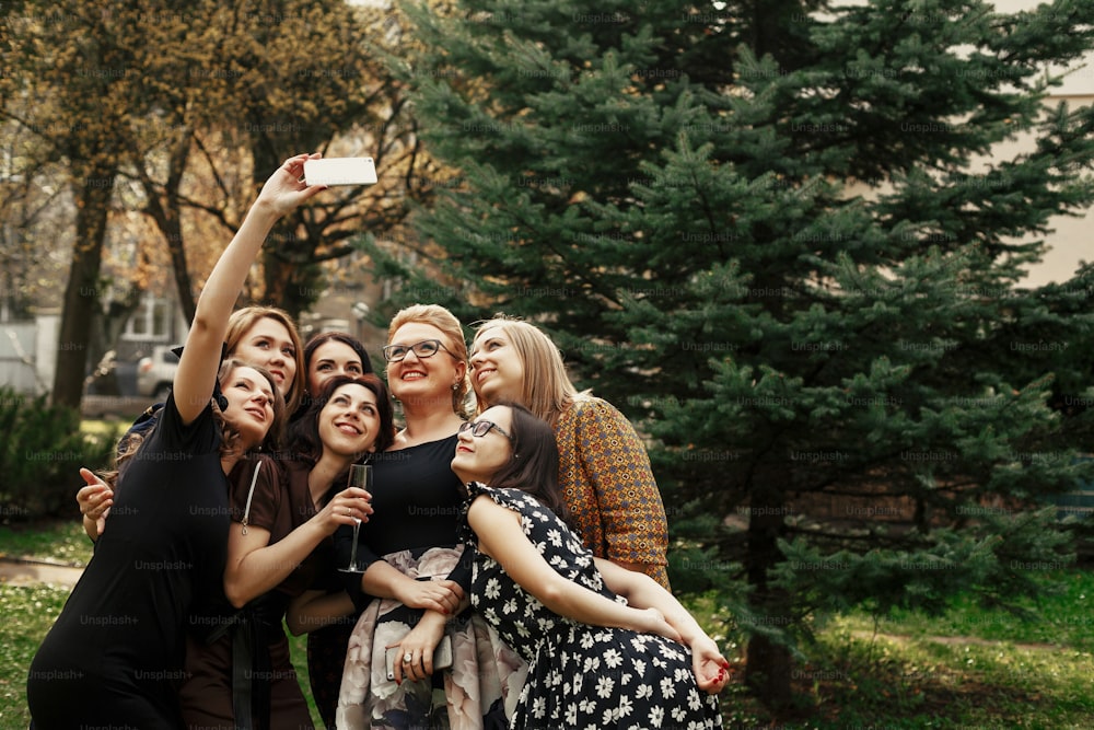 mujeres elegantes con estilo que toman selfies divertidas en la celebración soleada en el parque, concepto de vida feliz de lujo