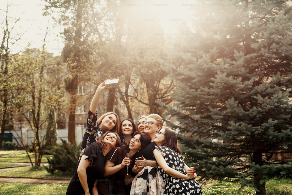 mujeres elegantes con estilo que toman selfies divertidas en la celebración soleada en el parque, concepto de vida feliz de lujo