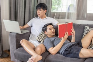 Pareja de hombre gay homosexual asiático que usa una computadora portátil que trabaja en casa con el novio leyendo un libro en el sofá de la sala de estar.
