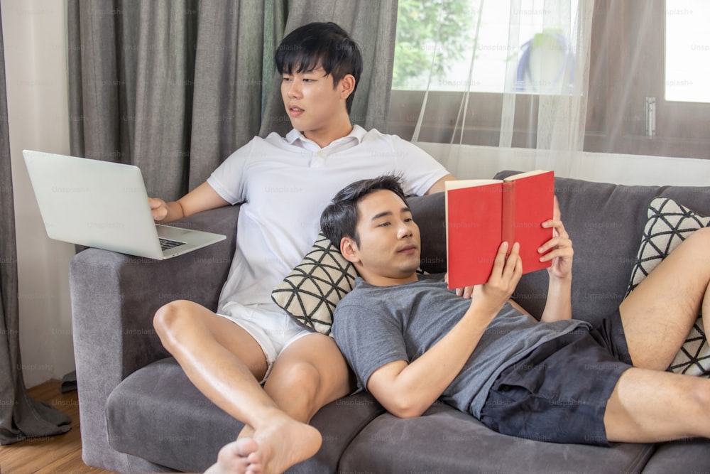Asian homosexual gay man couple using laptop working at home with boyfriend reading a book on sofa in living room.