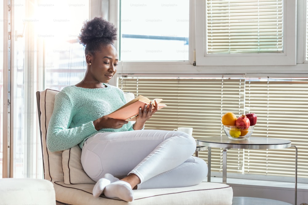 Cozy sofa and a beautiful girl, reading a book, concepts of home and comfort, place for text. Beautiful young black woman reading book near window at home. Stay home