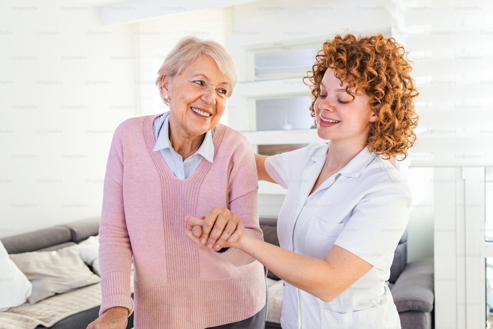 Smiling nurse helping senior lady to walk around the nursing home. Portrait of happy female caregiver and senior woman walking together at home. Professional caregiver taking care of elderly woman.