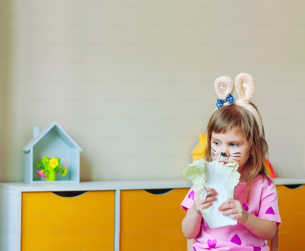 Adorable niña pequeña con pintura facial como conejito y orejas en la cabeza comiendo hoja de repollo sentada en la habitación de los niños.