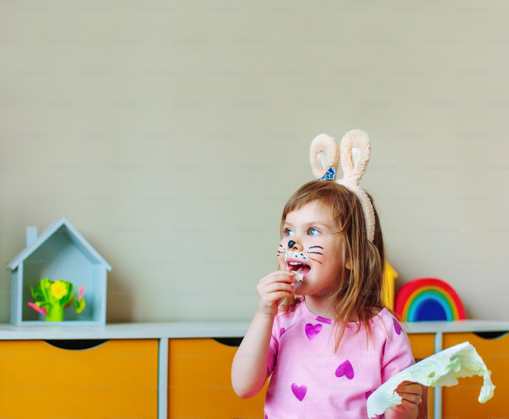 Adorabile bambina con il viso dipinto come un coniglietto e le orecchie sulla testa che mangia foglie di cavolo sedute nella stanza dei bambini.