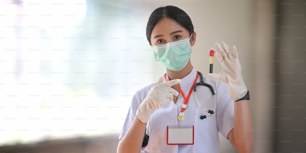 Ärztin mit medizinischer Maske und Handschuh, die einen Blutschlauch zum Test zeigt, während sie im modernen Untersuchungsraum über dem verschwommenen Krankenhaus als Hintergrund steht.