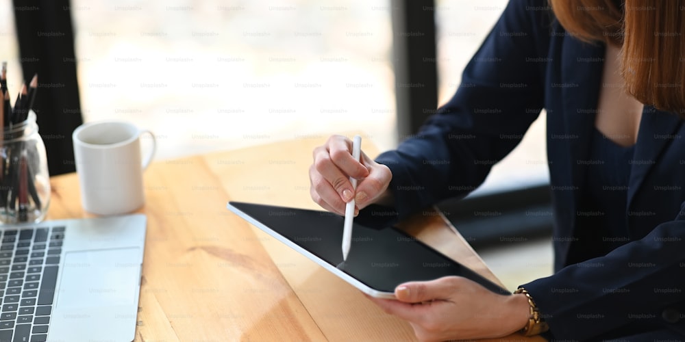 Abgeschnittenes Bild einer Geschäftsfrau, die einen Eingabestift hält, während sie ein Computer-Tablet benutzt und am hölzernen Schreibtisch über einem komfortablen Büro als Hintergrund sitzt.