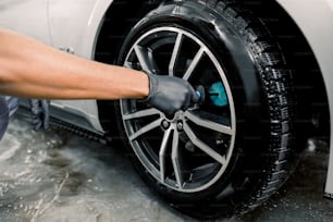 Servicio de lavado automático. Imagen de primer plano recortada de manos masculinas con guantes protectores negros, limpiando llantas de aleación de un automóvil de lujo con un cepillo especial para ruedas fundidas en un taller de detalles de vehículos.
