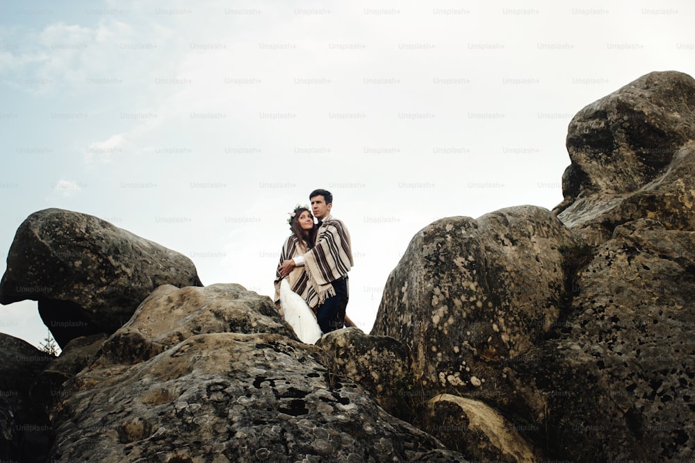 stylish gorgeous couple newlyweds hugging under rug on the rocks in the mountains in the evening light