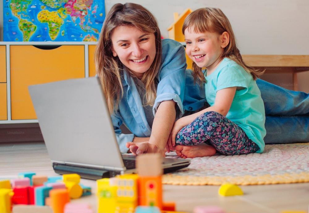 Smiling mother and toddler daughter looking at the notebook lying on the floor in the kids room. Distance online learning concept. Activities for kids during quarantine.