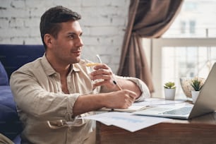 Attractive dreamy Caucasian middle-aged male freelancer holding a glass of immune-boosting beverage in his hand