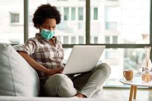 Black woman wearing face mask while working on a computer at home during virus epidemic.