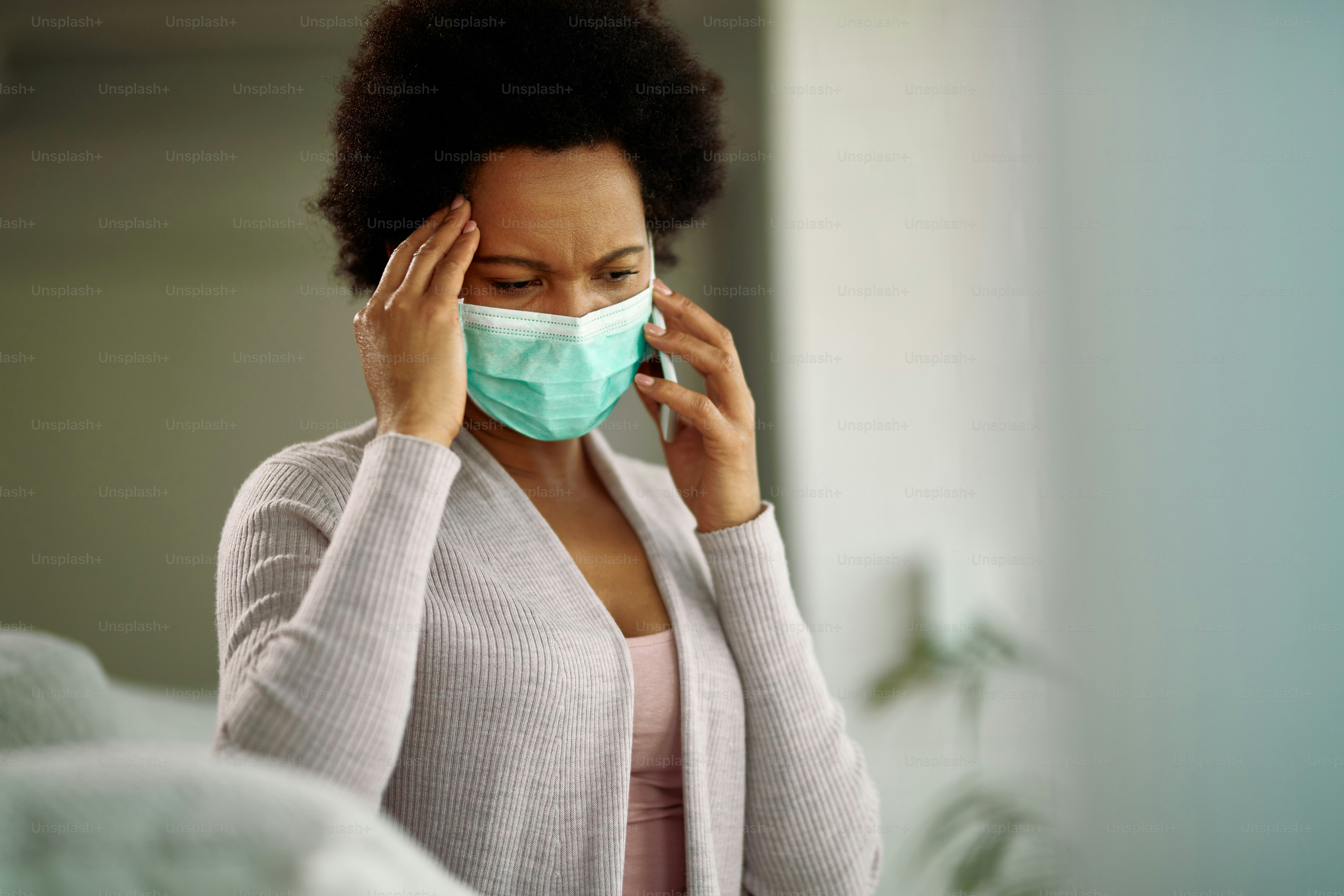 Worried African American woman wearing protective face mask while talking on mobile phone at home.