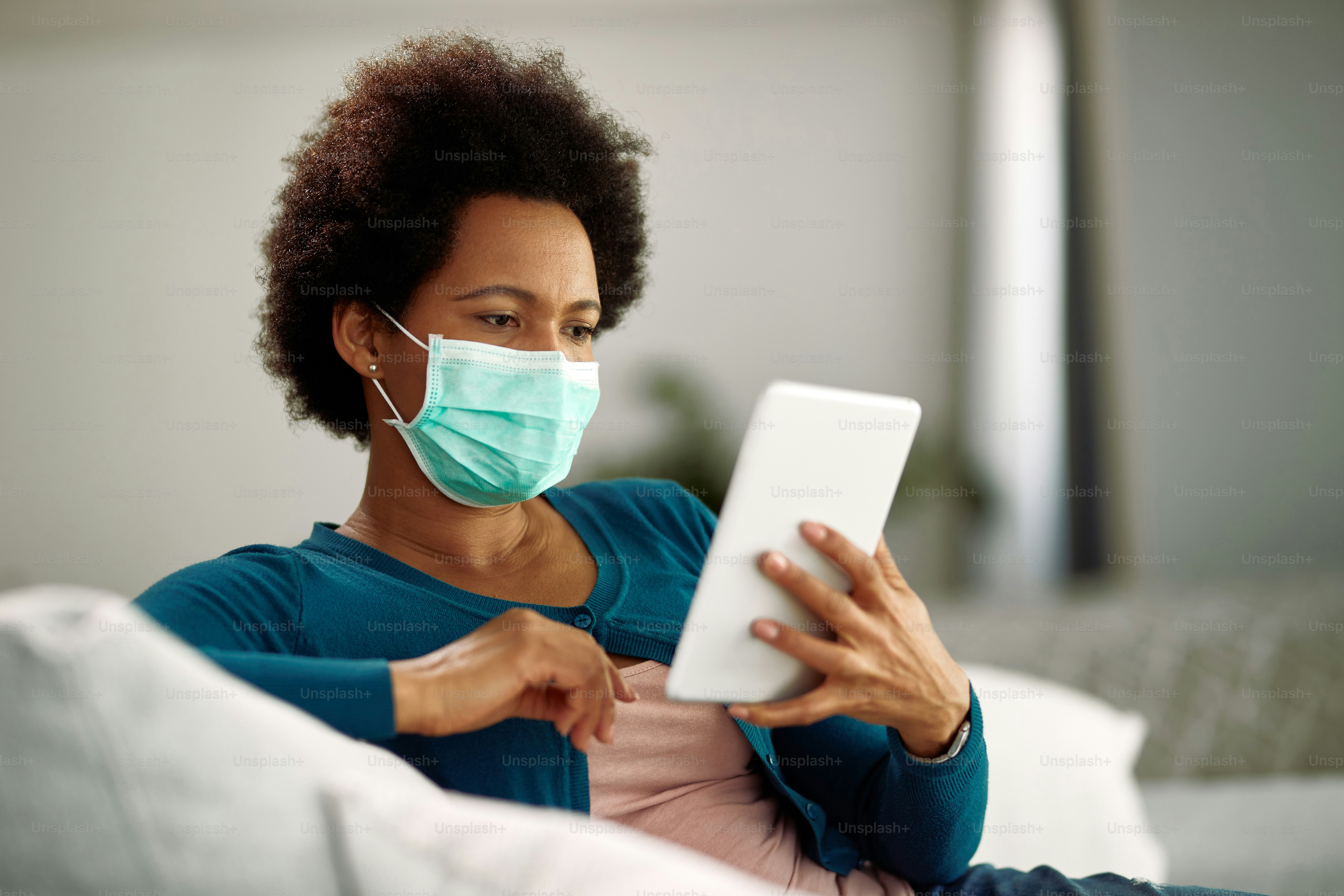 African American woman wearing face mask while surfing the net on touchpad in the living room.