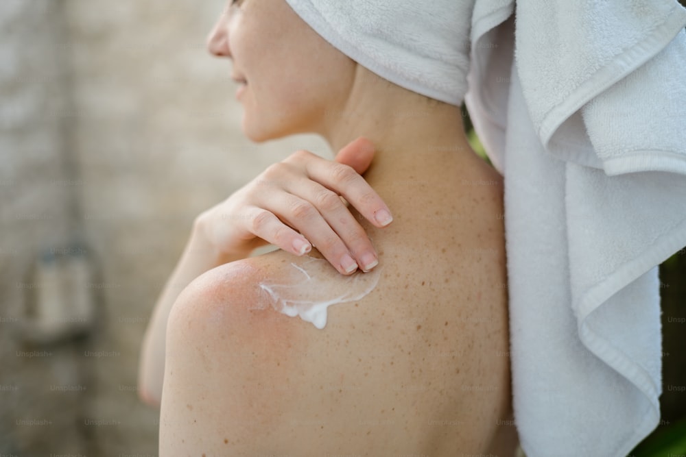Vue recadrée d’une jeune femme adulte debout dans la salle de bain, appliquant une crème hydratante sur l’épaule, faisant une procédure de soins de la peau