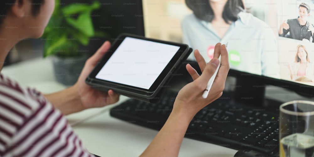 Image recadrée d’un homme créatif utilisant une tablette d’ordinateur avec un écran blanc et un stylet tout en faisant une vidéoconférence et assis devant son écran d’ordinateur au bureau de travail.