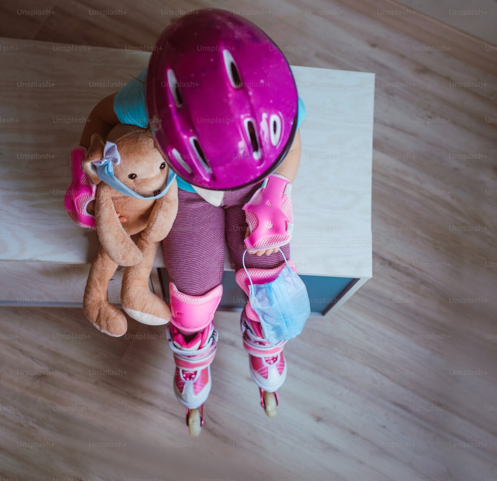 Top view of little girl wearing pink roller skates, knee pads, wristbands and helmet holding her face mask and rabbit soft toy in the mask sitting indoor. Social distance stay at home during Covid-19 Pandemic concept. Selective focus on the mask.