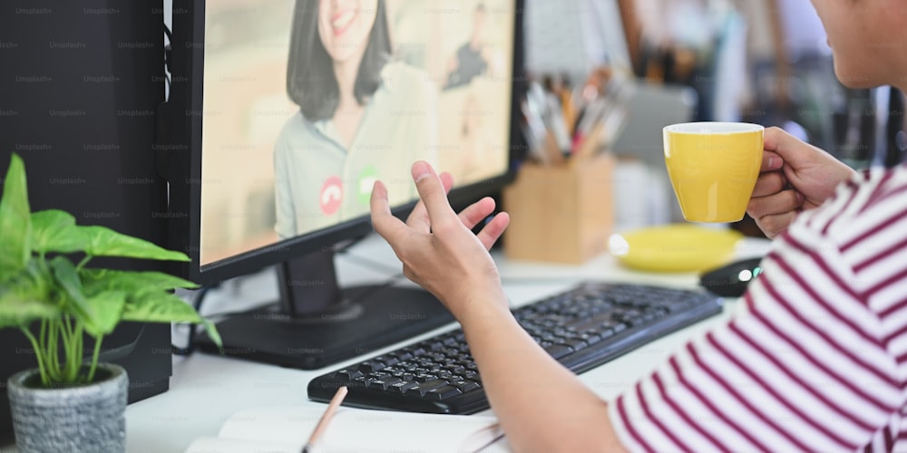 Cropped image of creative man holding coffee cup and making a video conference with his colleague while sitting at the white working desk over modern living room as background. Remote working concept.