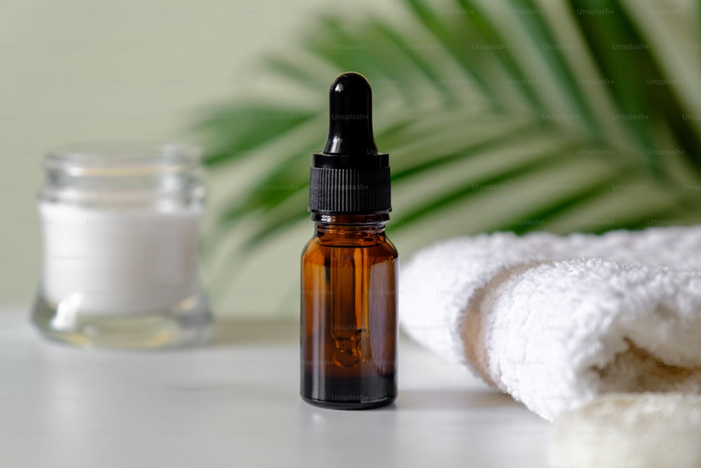 Serum or essential oil bottle with moisturizer and towel, palm leaf on the background. Clear amber glass dropper bottle with natural organic cosmetic, SPA products, skin treatment, body care concept.
