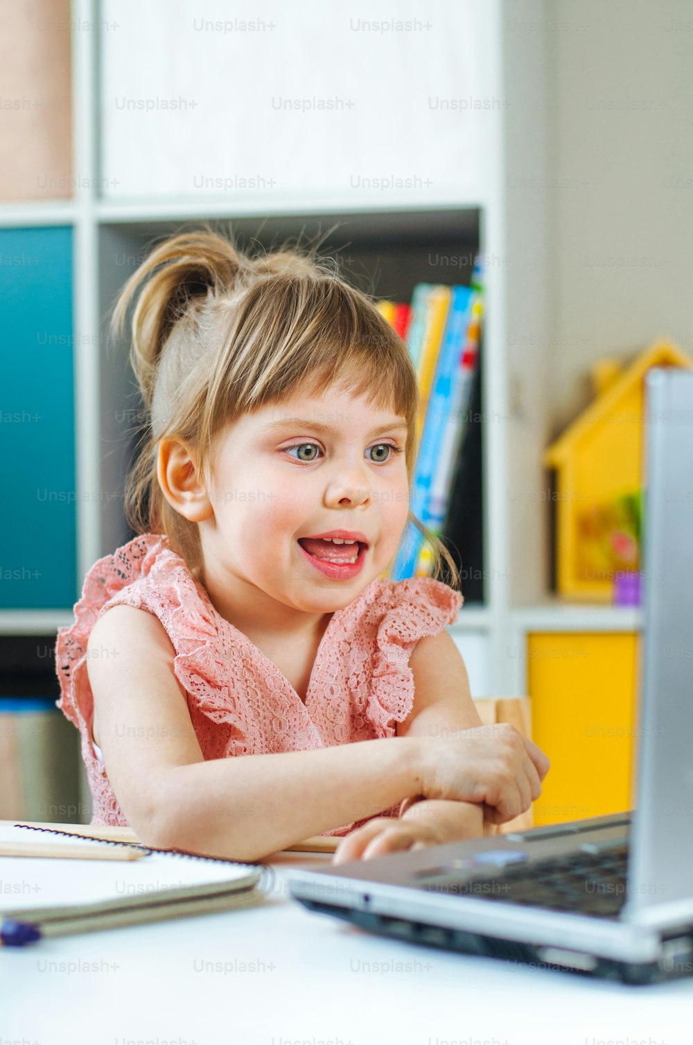 Happy little toddler girl using notebook for distance learning sitting at the table in the kids room. Distance online learning concept.