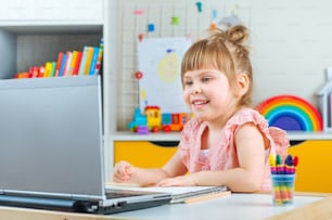 Smiling little girl using notebook for distance learning sitting at the table in the kids bedroom. Distance online learning concept.