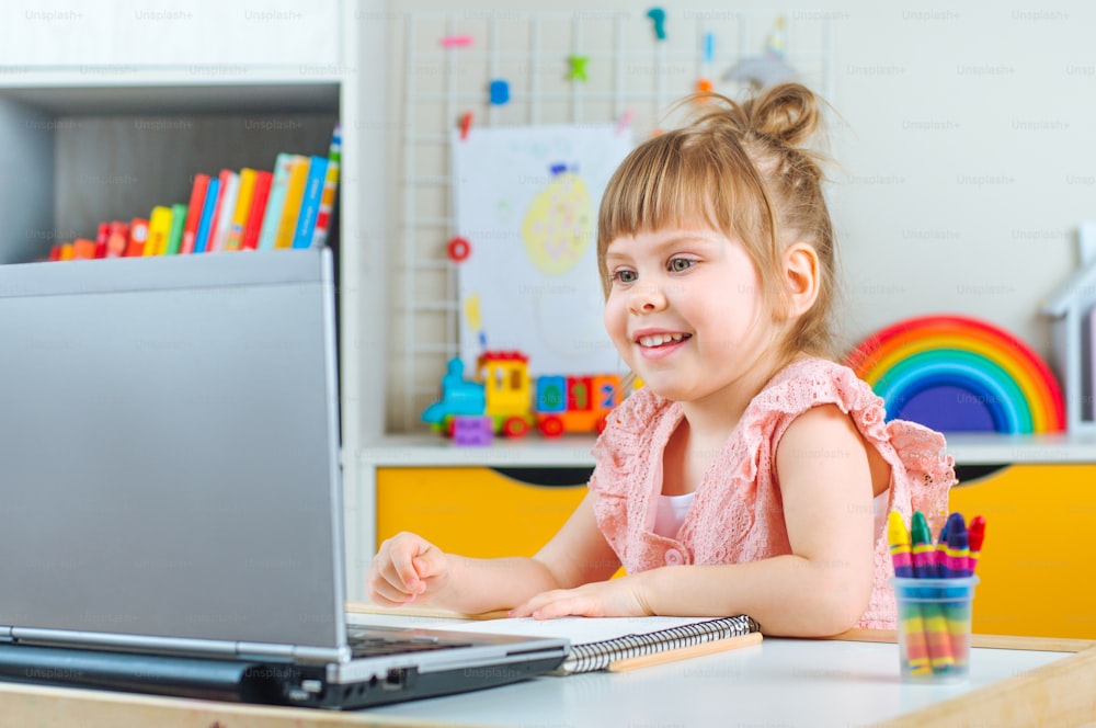 Smiling little girl using notebook for distance learning sitting at the table in the kids bedroom. Distance online learning concept.