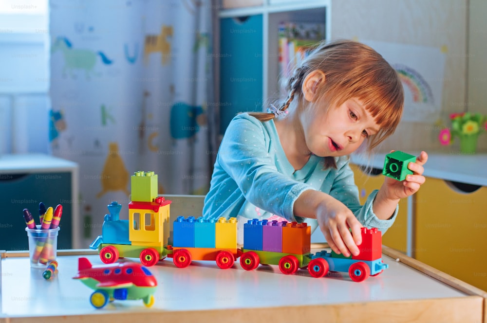 子供部屋でカラフルな立方体で子供のプラスチック製のおもちゃの列車で遊んでいる小さな女の子。子供のためのホームアクティビティ。